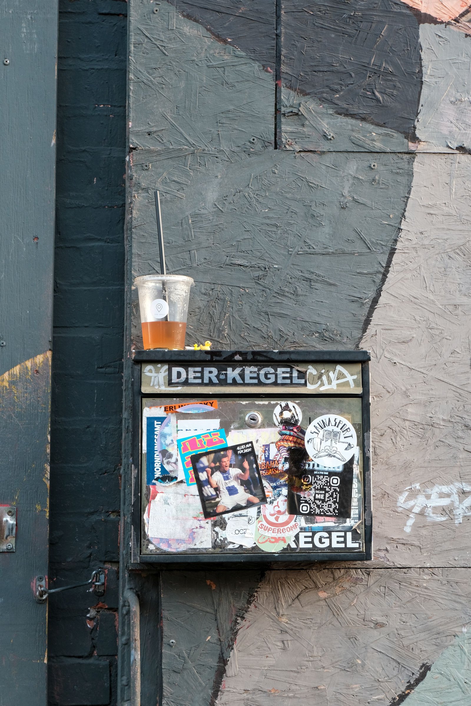 Beklebter Kasten an einer bemalten Holzwand, darauf ein halbvoller Becher